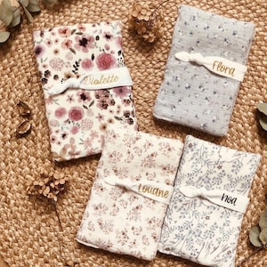 Puede incluir: Cuatro mantas para beb&eacute; con estampados florales en tonos rosa, morado y azul. Cada manta est&aacute; asegurada con una banda blanca bordada con un nombre: Violette, Flora, Louane y Noa. Las mantas est&aacute;n dispuestas sobre una superficie tejida.