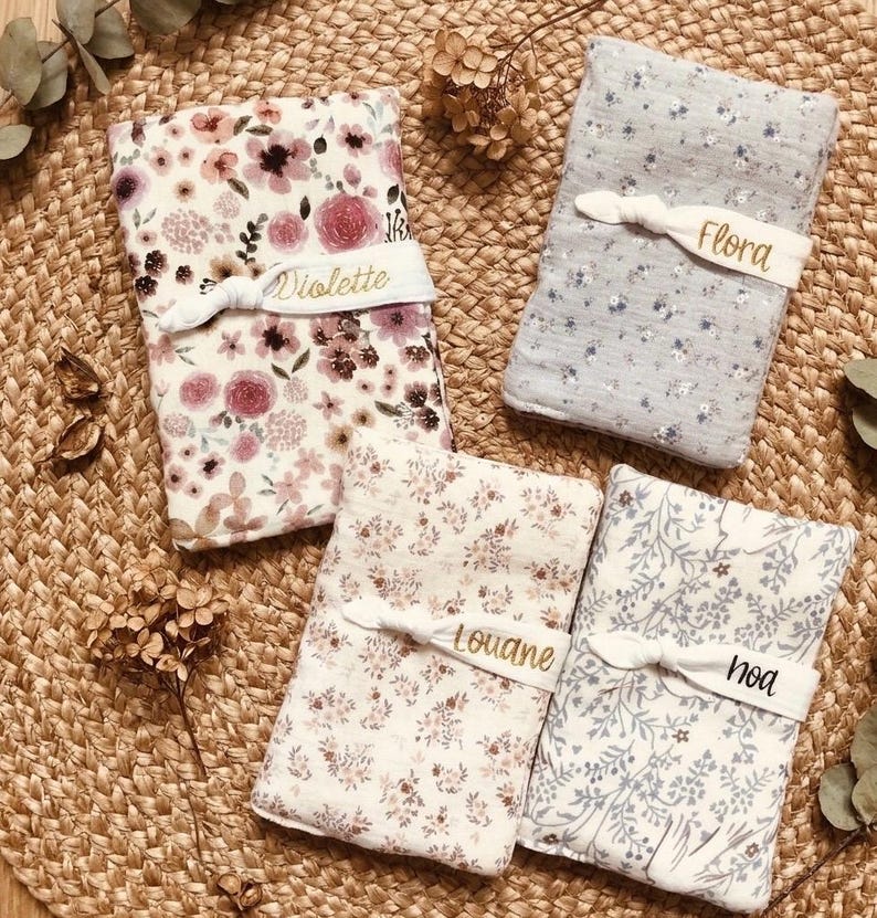 Puede incluir: Cuatro mantas para beb&eacute; con estampados florales en tonos rosa, morado y azul. Cada manta est&aacute; asegurada con una banda blanca bordada con un nombre: Violette, Flora, Louane y Noa. Las mantas est&aacute;n dispuestas sobre una superficie tejida.