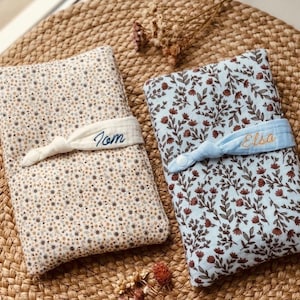 Puede incluir: Dos cambiadores para beb&eacute;s. Uno es beige con un estampado floral y el nombre "Tom" bordado en una banda blanca. El otro es azul claro con un estampado floral y el nombre "Elsa" bordado en una banda azul claro.