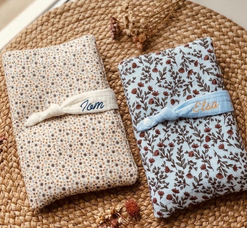 Puede incluir: Dos cambiadores para beb&eacute;s. Uno es beige con un estampado floral y el nombre "Tom" bordado en una banda blanca. El otro es azul claro con un estampado floral y el nombre "Elsa" bordado en una banda azul claro.
