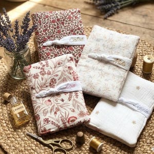 Puede incluir: Cuatro cuadernos cubiertos de tela con estampados florales y bandas el&aacute;sticas bordadas con nombres. Tambi&eacute;n se ven una peque&ntilde;a botella de aceite de lavanda, unas tijeras y un tarro de ramitas de lavanda. Los cuadernos son en tonos rojos, crema y blanco.