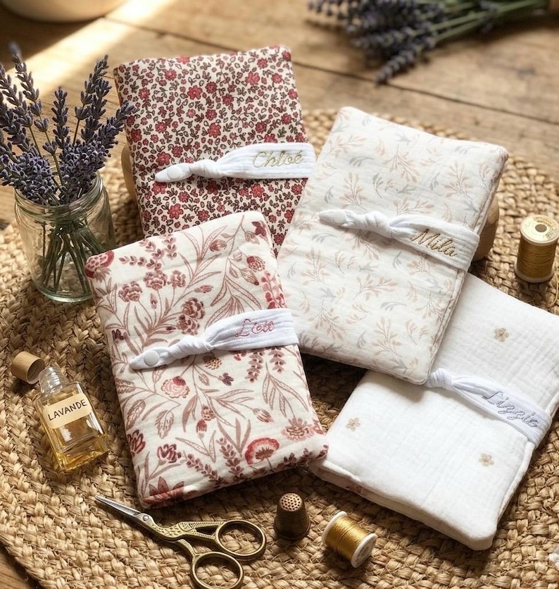 Puede incluir: Cuatro cuadernos cubiertos de tela con estampados florales y bandas el&aacute;sticas bordadas con nombres. Tambi&eacute;n se ven una peque&ntilde;a botella de aceite de lavanda, unas tijeras y un tarro de ramitas de lavanda. Los cuadernos son en tonos rojos, crema y blanco.