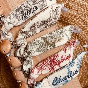 May include: Five fabric pacifier clips featuring floral patterns in various colors, each embroidered with a name: Nino, Alma, Jade, Fabien, and Charlie. Each clip includes a round wooden fastener and a small white loop for attachment.
