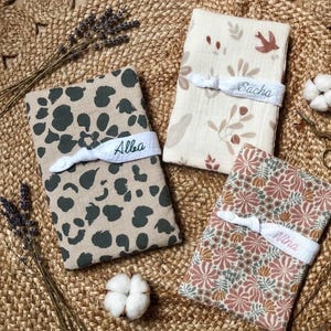 Puede incluir: Tres mantas de beb&eacute; personalizadas. Una tiene un estampado de animales en color beige y verde oscuro, otra tiene un estampado floral en rosa, verde y marr&oacute;n, y la tercera tiene un dise&ntilde;o floral y de p&aacute;jaros. Cada manta est&aacute; envuelta con una banda blanca bordada con un nombre.