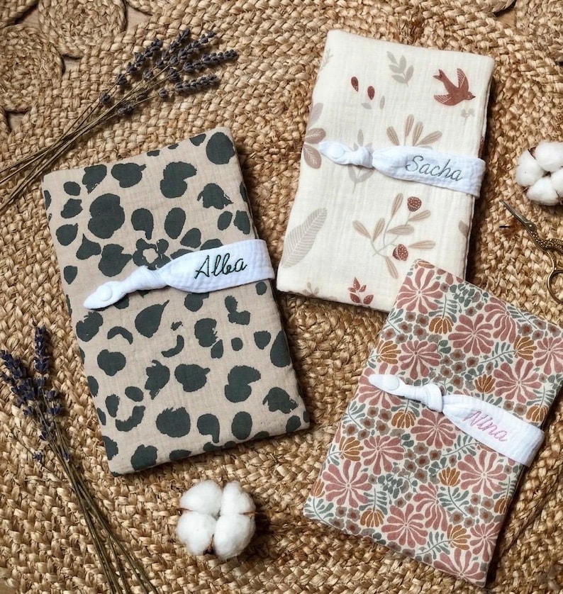 Puede incluir: Tres mantas de beb&eacute; personalizadas. Una tiene un estampado de animales en color beige y verde oscuro, otra tiene un estampado floral en rosa, verde y marr&oacute;n, y la tercera tiene un dise&ntilde;o floral y de p&aacute;jaros. Cada manta est&aacute; envuelta con una banda blanca bordada con un nombre.