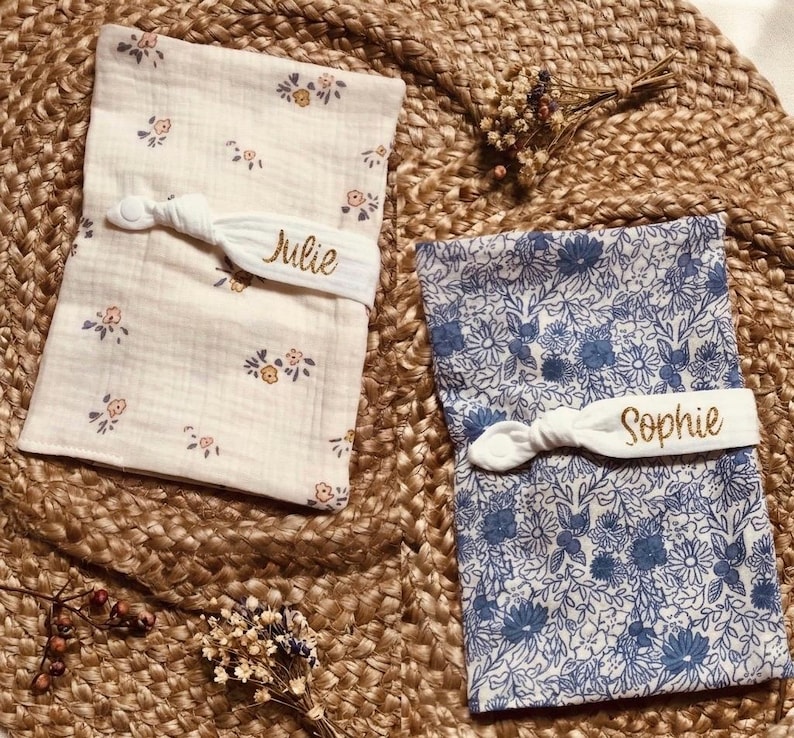 Puede incluir: Dos baberos de tela para beb&eacute;, uno crema con estampado floral y otro azul con un estampado floral. Cada babero se asegura con una banda el&aacute;stica blanca bordada con un nombre, "Julie" y "Sophie".