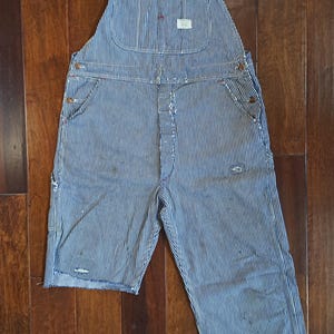 May include: Blue and white striped denim overalls with adjustable straps and multiple pockets. The overalls have a classic workwear design, featuring a bib front and a relaxed fit. The fabric shows signs of wear and tear, adding to its vintage appeal.
