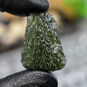 Peut inclure: Une tektite de couleur vert foncé, de forme irrégulière et de surface rugueuse.