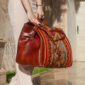 Peut inclure: Un grand sac de voyage en cuir marron avec un dessus en tissu à motifs colorés. Le sac a un design structuré avec une poignée supérieure et une bandoulière. Le tissu présente des motifs géométriques rouges, jaunes et bleus. Le sac est tenu par une personne.