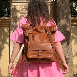May include: A brown leather backpack with a flap closure and adjustable straps. The backpack has a large main compartment and a smaller front pocket. The backpack is worn by a person in a pink dress.