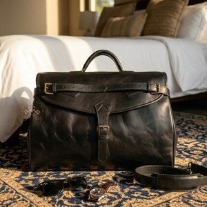 May include: A black leather briefcase featuring a top handle, a horizontal strap with a buckle, and a central vertical strap with a buckle closure. A detachable shoulder strap, a wristwatch, and sunglasses are placed in front of the bag.