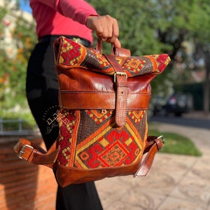 以下が含まれることがあります： カラフルな織り生地のフラップが付いた茶色の革のバックパック。バックパックはバックル留めと調節可能なストラップが付いています。