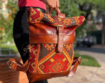 Puede incluir: Una mochila de cuero marrón con una solapa de tejido tejido de colores. La mochila tiene un cierre de hebilla y correas ajustables.