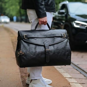 May include: A large black leather briefcase featuring a top handle, a horizontal strap with a buckle across the front flap, and a vertical central strap with a buckle closure. The bag has a structured rectangular shape with visible stitching details.