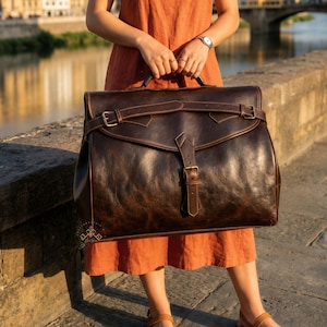 May include: A large, dark brown leather satchel featuring a wide, horizontal strap across the top flap, secured by a central vertical buckle closure. The bag has a smooth, distressed finish with visible stitching and antique-style metal hardware.