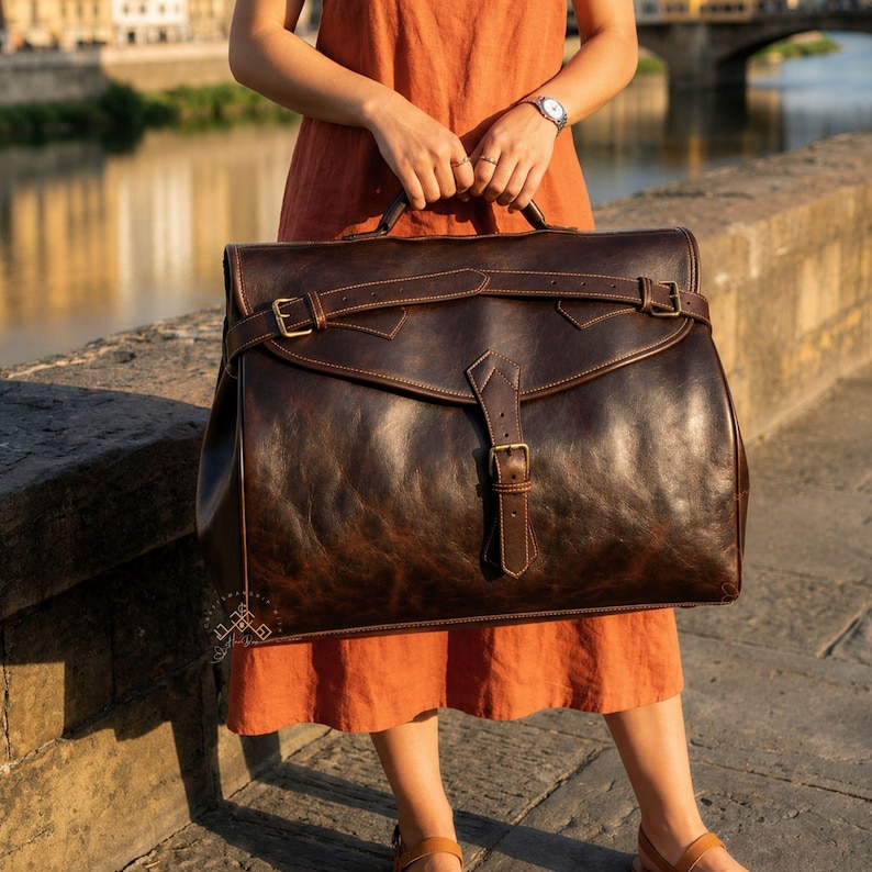 May include: A large, dark brown leather satchel featuring a wide, horizontal strap across the top flap, secured by a central vertical buckle closure. The bag has a smooth, distressed finish with visible stitching and antique-style metal hardware.