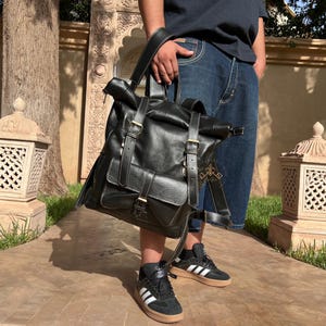May include: A black leather backpack with multiple buckles and straps. The backpack has a flap that folds over the top and is secured with a buckle. The backpack is being worn by a person.