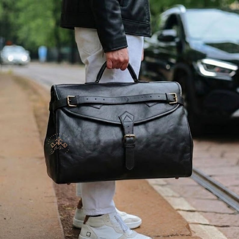 May include: A large black leather briefcase featuring a top handle, a horizontal strap with a buckle across the front flap, and a vertical central strap with a buckle closure. The bag has a structured rectangular shape with visible stitching details.