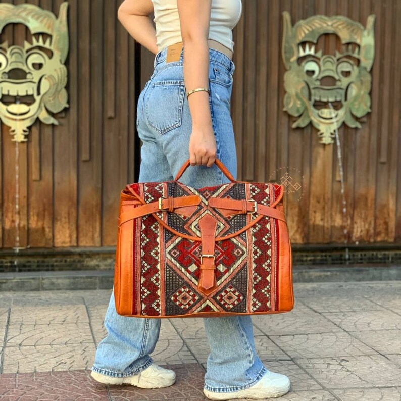 May include: A large, brown leather travel bag with a woven tapestry design in shades of red, blue, and grey. The bag has two buckles and a strap.