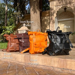 May include: Four leather backpacks in various colors: brown (almond), dark brown (cafe), orange (havan), and black. Each backpack features a roll-top design, adjustable straps, and multiple pockets. The bags are displayed outdoors.