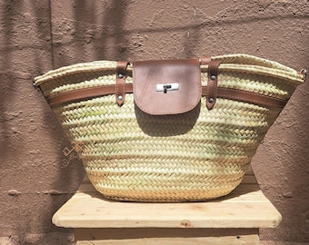 Puede incluir: Bolso tote de paja tejida con detalles de cuero marrón. El bolso tiene una solapa de cuero marrón con un cierre plateado. El bolso está sobre una superficie de madera.
