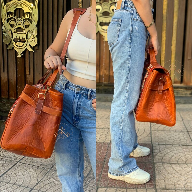 May include: A brown leather tote bag with a buckle closure and a long strap. The bag is being worn by a person wearing blue jeans and a white tank top.