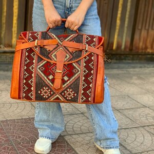 May include: A large, brown leather travel bag with a red, white and black geometric pattern. The bag has a flap closure with a buckle and a strap.