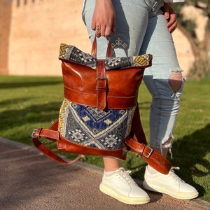 Puede incluir: Una mochila de cuero marr&oacute;n con una solapa de tejido tejido de colores. La mochila tiene un cierre de hebilla y correas ajustables.