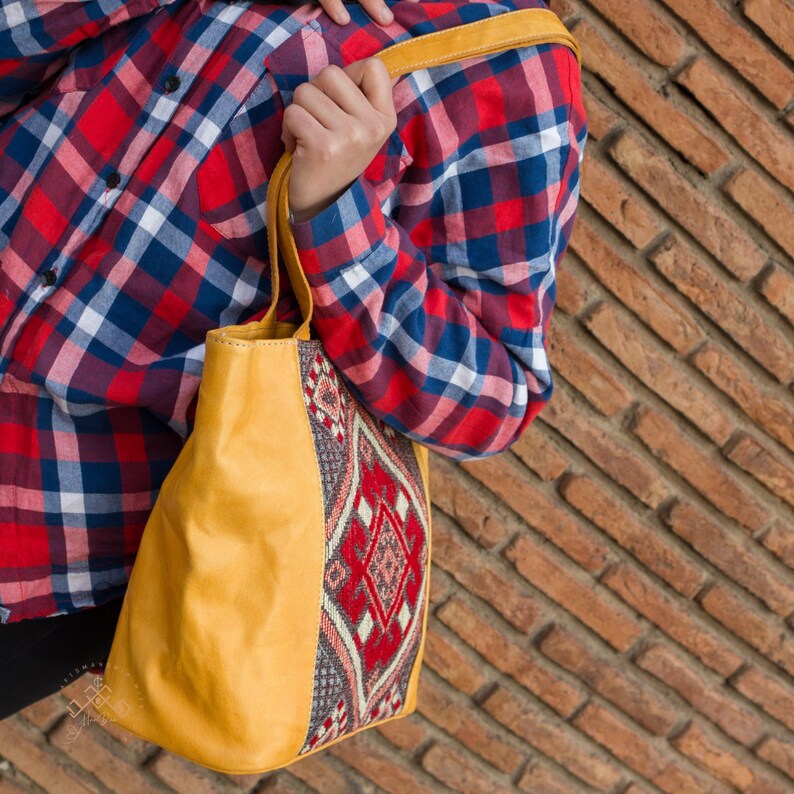 May include: A yellow leather tote bag with a colorful woven fabric panel featuring a geometric pattern. The bag has a top handle and a shoulder strap.