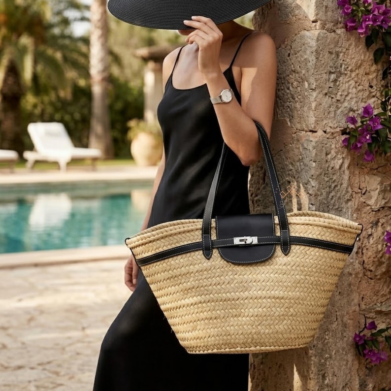 May include: A woven straw tote bag with black leather detailing and a black flap closure. The bag features black handles and a silver-coloured clasp. The bag is tan and black, suitable for a beach or summer outing.