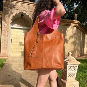 May include: A large, brown leather tote bag with a tassel hanging from the zipper pull. The bag has a wide, open top and a single strap.