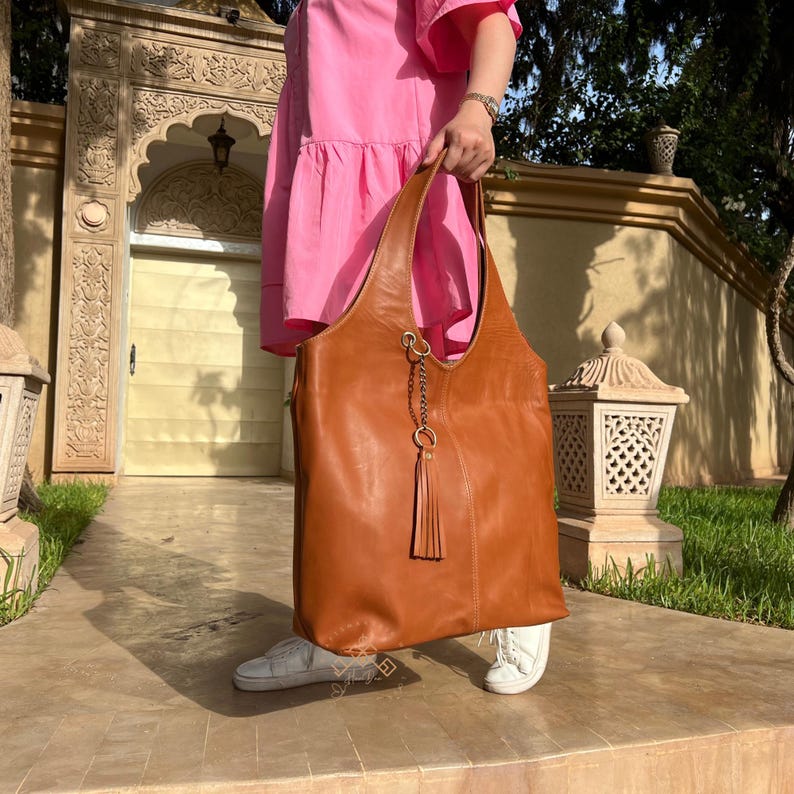 May include: A large, brown leather tote bag with a long tassel hanging from the front. The bag has a single strap and a metal ring with a chain attached to it.