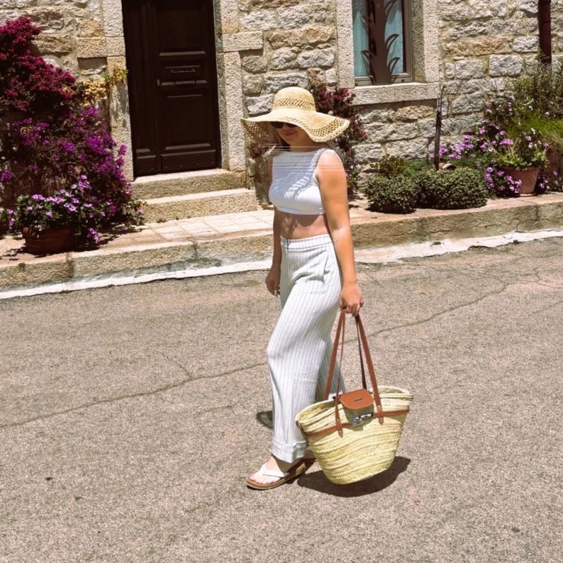 May include: A woman in a straw hat, white crop top, and striped trousers, carrying a straw tote bag with brown leather handles. She is wearing sandals. The background shows a stone building and flowers.