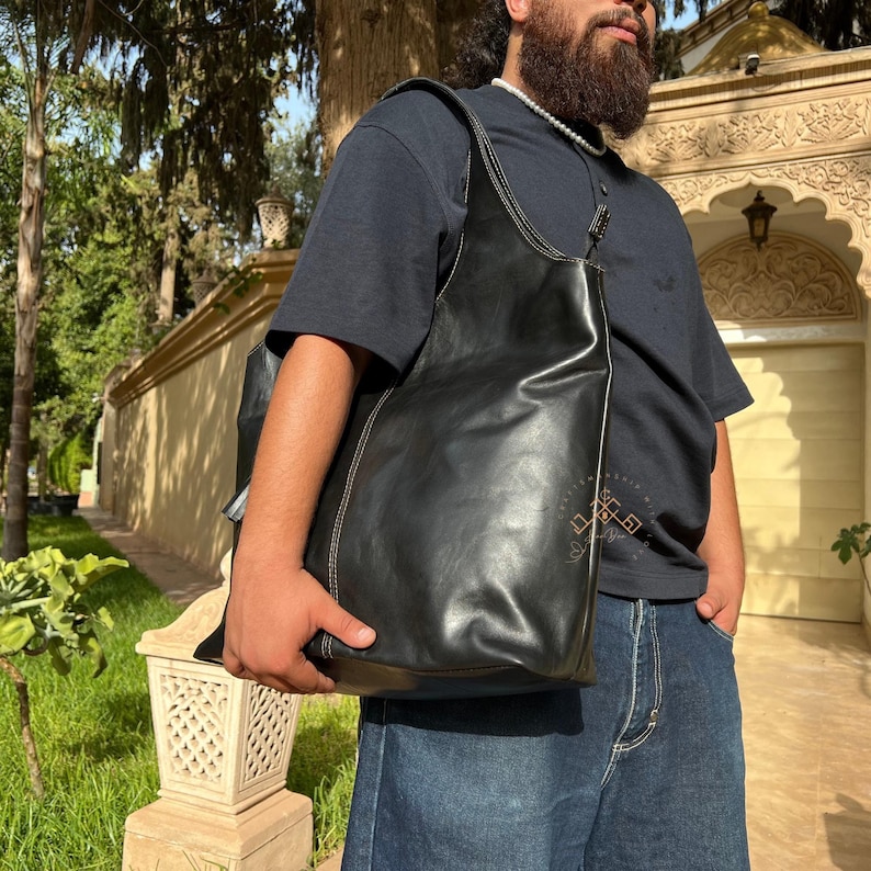 May include: A black leather tote bag with a white stitching detail. The bag is being held by a person wearing a dark blue t-shirt and blue jeans.