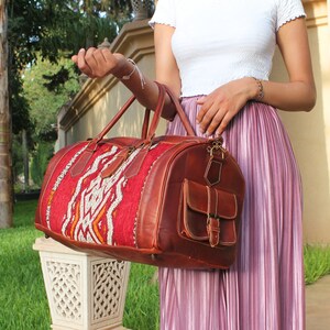 May include: A brown leather duffel bag with a red and white woven pattern on the front. The bag has a large front pocket with a buckle closure.