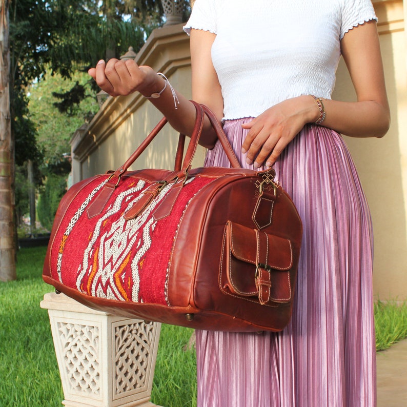 May include: A brown leather duffel bag with a red and white woven pattern on the front. The bag has a large front pocket with a buckle closure.