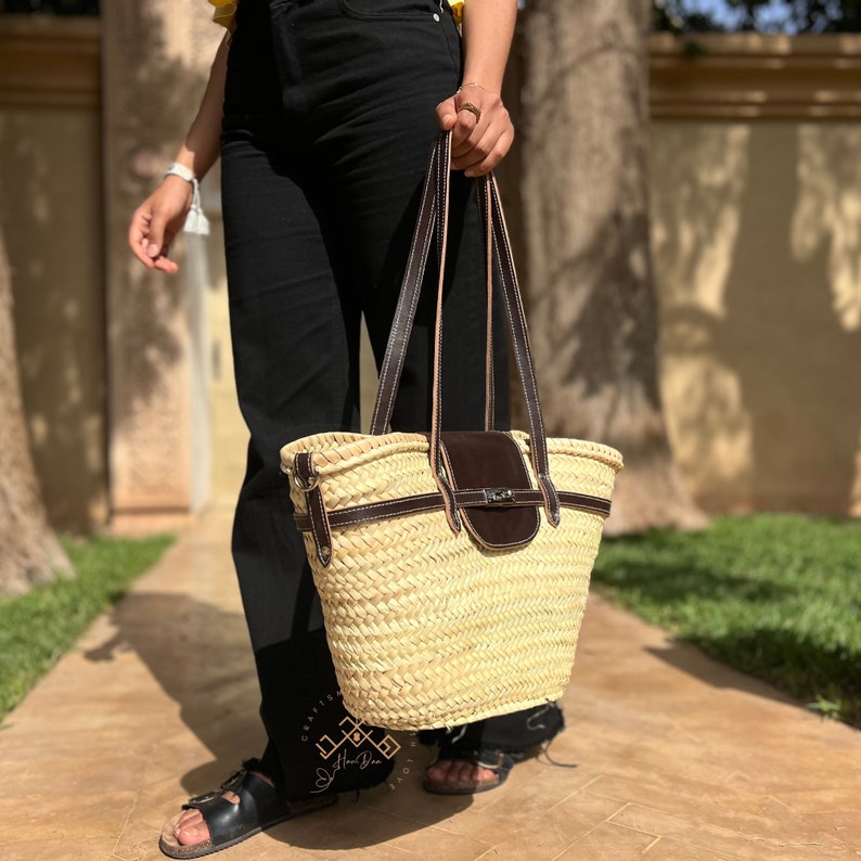 May include: A natural woven straw tote bag with brown leather detailing and a flap closure. The bag features long leather straps. The bag is held by a person wearing black trousers and sandals.