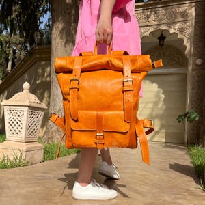 May include: A large, orange leather backpack with a roll-top closure and multiple buckles. The backpack is made of a distressed leather and has a vintage look.