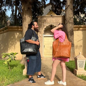 May include: Two people are standing outside, each carrying a large leather tote bag. The person on the left is wearing a black shirt and blue jeans, and the person on the right is wearing a pink shirt and shorts. Both bags have a tassel hanging from the handle.