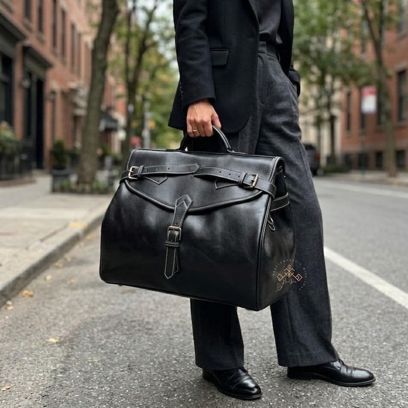 May include: A large black leather travel bag featuring a structured rectangular shape, a top handle, and a front flap secured with two horizontal buckled straps and a central vertical buckled strap. The bag has visible stitching and metal hardware.