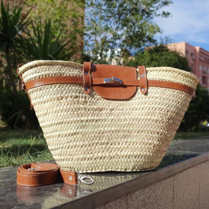 May include: Woven straw tote bag with brown leather detailing. Includes a brown leather flap with a silver clasp and a detachable brown leather shoulder strap. The bag has a wide opening and a robust base.