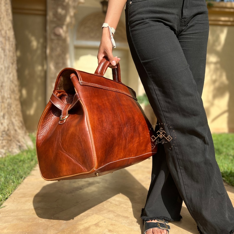 May include: A large, structured brown leather doctor-style bag with a top handle, brass-toned hardware, and visible stitching. The bag features a wide, hinged opening, a smooth finish with natural grain variations, and a logo embossed on the side.