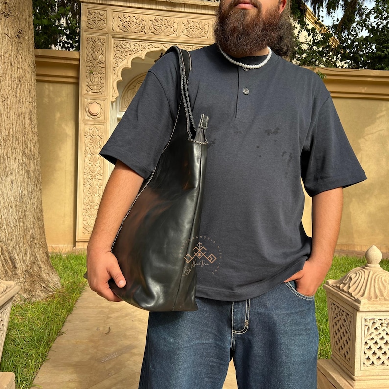 May include: A person wearing a dark blue short-sleeved shirt with a black leather tote bag. The shirt has a mock neck and a single button closure. The person is wearing blue jeans.