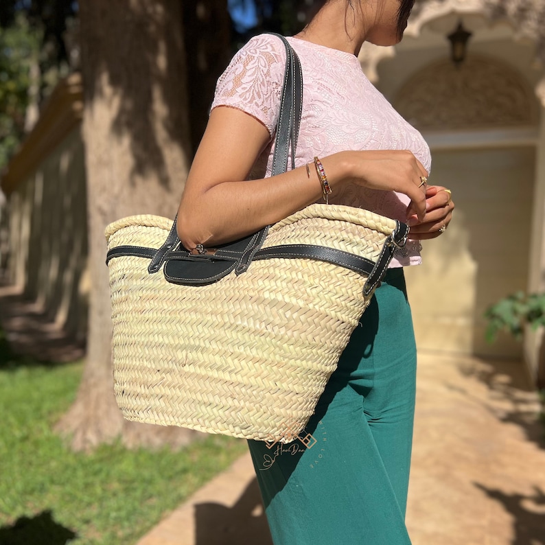 May include: A natural woven straw tote bag with black leather handles and trim. The bag is rectangular and is carried by a person wearing a pink lace top and green trousers. The bag has a woven pattern.