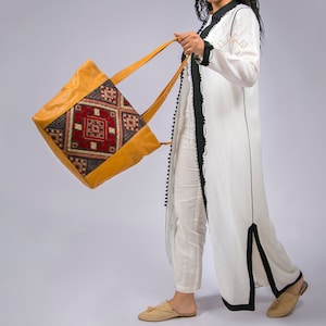 May include: A woman wearing a white dress with black trim carries a large, yellow leather tote bag with a colorful geometric pattern.