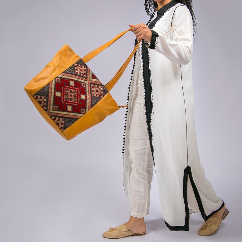 May include: A woman wearing a white dress with black trim carries a large, yellow leather tote bag with a colorful geometric pattern.
