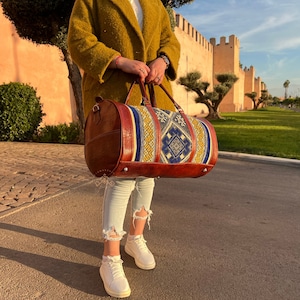 Puede incluir: Una bolsa de viaje de cuero marr&oacute;n con un patr&oacute;n tejido colorido. La bolsa tiene un asa superior y un cierre de cremallera.