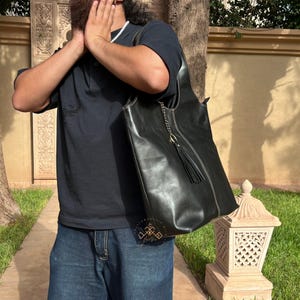May include: A large black leather tote bag with a tassel and a gold chain detail. The bag is being held by a person wearing a black shirt and blue jeans.