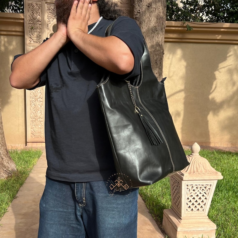 May include: A large black leather tote bag with a tassel and a gold chain detail. The bag is being held by a person wearing a black shirt and blue jeans.