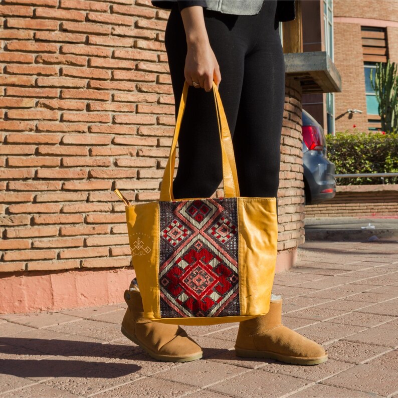 May include: A yellow leather tote bag with a colorful embroidered panel featuring geometric patterns. The bag has a top handle and a wide opening.